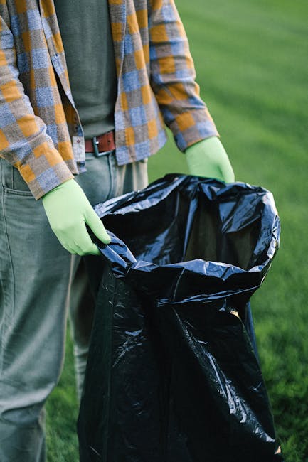 A person standing outdoors on a grassy area is holding a large black rubbish bag open, preparing for waste disposal. The individual is wearing a long-sleeved green shirt, a plaid jacket with shades of brown, blue, and yellow, and green gloves for handling waste. The background shows a well-maintained lawn with lush green grass. The scene relates to private waste handling or rubbish clearance, emphasizing independent collection methods characteristic of rubbish removal services. The focus is on the manual loading or preparation phase of rubbish disposal, with the person's hand visible gripping the edge of the black plastic bag and the bag itself appearing empty but ready to be filled with household or garden waste. The setting suggests an organized approach to waste collection, aligned with services provided by companies such as Rubbish Clearance Sutton, supporting onsite clearance or alternative waste disposal options outside municipal collection systems.