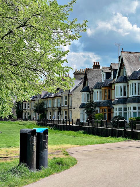 A row of traditional terraced houses with bay windows and slate roofs situated behind a black metal fence. In the foreground, there are two black waste bins positioned near a grassy area and a paved pathway. To the left, a large leafy tree with light green foliage extends over the scene, casting some shade. The background shows a partly cloudy sky with patches of blue, contributing to a bright and clear daytime setting. The scene appears to depict an urban residential street, where the presence of waste bins suggests regular rubbish collection. This setting aligns with the context of private waste handling or rubbish clearance services often seen in urban environments, supporting the theme of rubbish removal and disposal options.