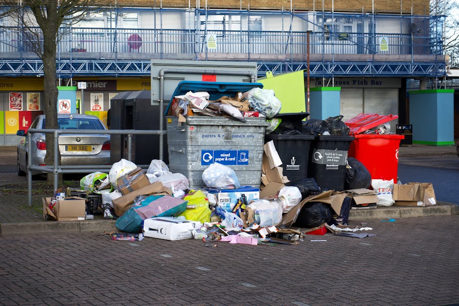 A large pile of mixed household waste and recyclable materials accumulated on a paved area beside a street, featuring assorted cardboard boxes, plastic bags, paper, and packaging supplies spilling over from overflowing black and red bins into the surrounding space. The black recycling bin with a blue lid, designated for mixed paper and card, stands prominently in the centre, filled to capacity with various papers and cardboard protruding from the top. To its right, a black bin with a dedicated disposal label and a red bin are also overflowing with rubbish. Surrounding the waste are scattered boxes, plastic bottles, and miscellaneous packaging, indicating recent or ongoing collection activity. In the background, there is an urban environment with a building under construction or refurbishment, covered in scaffolding and netting, and a row of shopfronts with storefront signage. The scene is well-lit, with natural daylight highlighting the details of the waste materials and the urban surroundings, illustrating an example of private waste handling or on-site rubbish clearance potentially managed by a service like Rubbish Clearance Sutton.